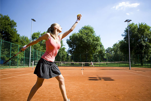Female tennis player preparing to serve on a sunny outdoor clay court surrounded by green trees.