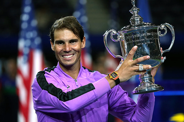 Rafael Nadal holding the US Open trophy, smiling in a purple jacket with an American flag in the background.