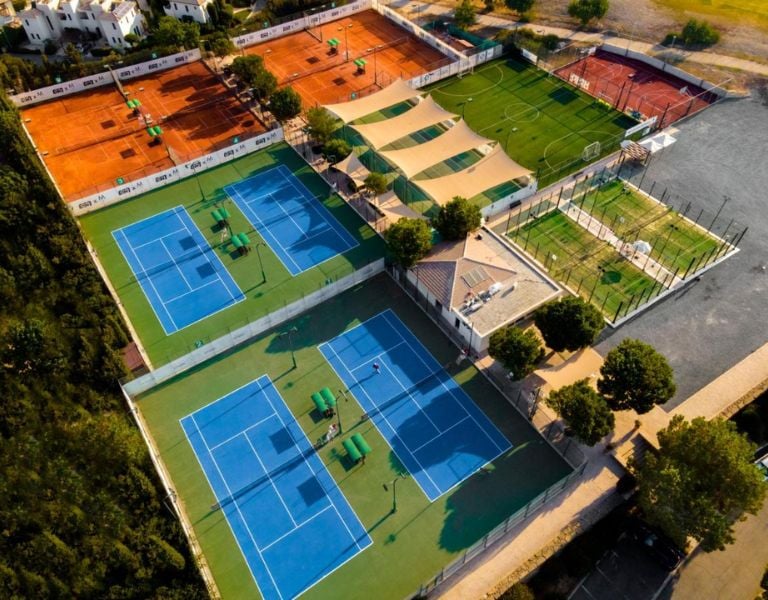 Aerial view of Aphrodite Hills sports courts including blue and green tennis courts, clay courts, and adjacent greenery.