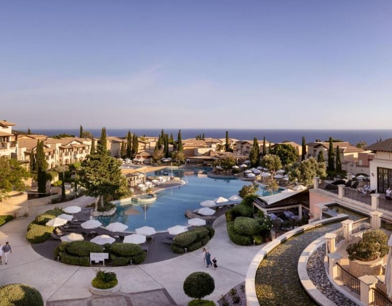 A scenic view of the Aphrodite Hills Resort pool area with umbrellas and villas, set against the Mediterranean landscape and coastline.