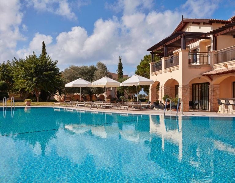 Luxury villa at Aphrodite Hills Resort with a pool, sun loungers, and white umbrellas under a clear blue sky.