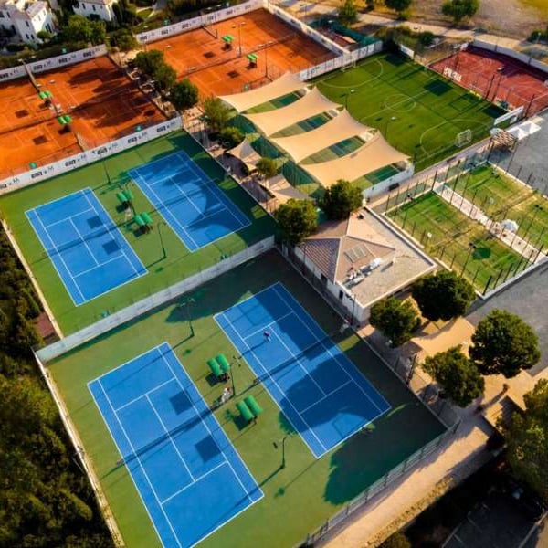 Aerial view of Aphrodite Hills Resort tennis courts, including blue hard courts and orange clay courts, surrounded by green fields and trees.