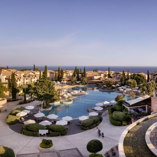 Aerial view of Aphrodite Hills Resort with a large pool, sun loungers, Mediterranean-style buildings, and a view of the sea.