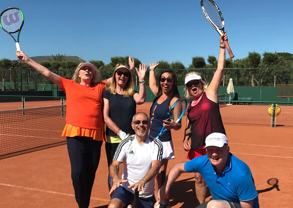 Group of excited tennis players holding rackets on a clay court with a clear sky and trees in the background, at the Active Away 2022 event.