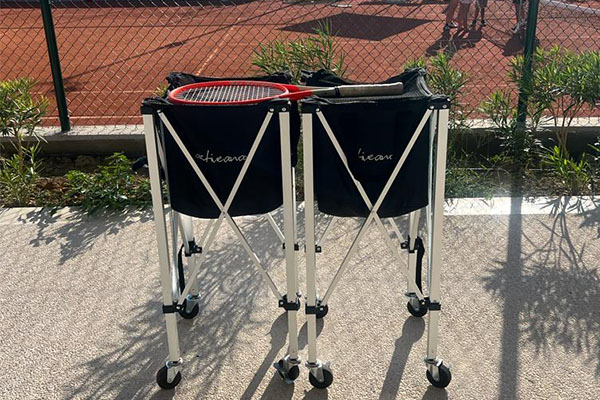 Two Active Away tennis ball baskets on court with wheels for mobility, accompanied by a tennis racket on top, on a sunlit clay court.