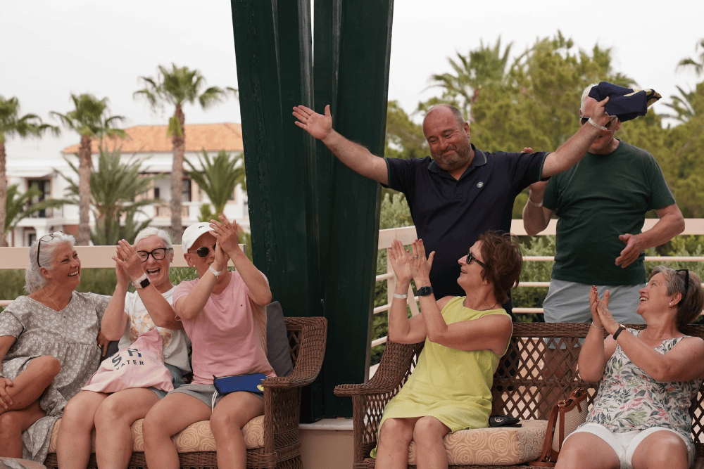Group of happy individuals clapping and enjoying a tennis holiday experience with Active Away, seated outdoors on wicker chairs.