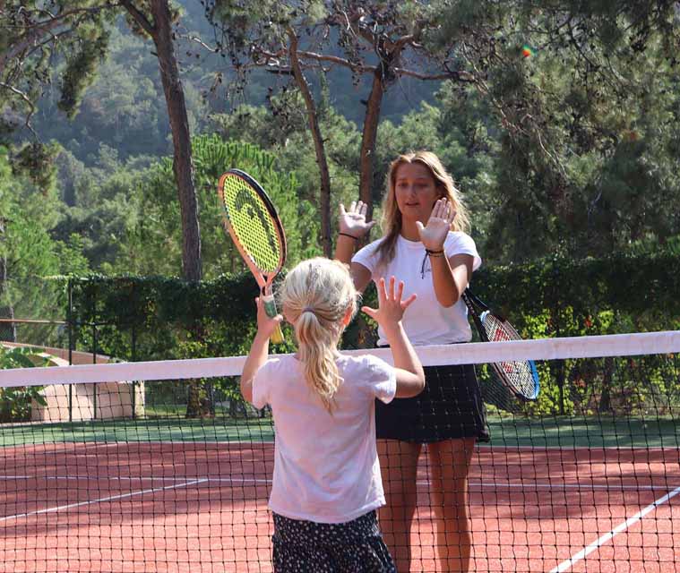 Woman and young girl playing tennis on an outdoor court, surrounded by trees, as they exchange high-fives over the net.
