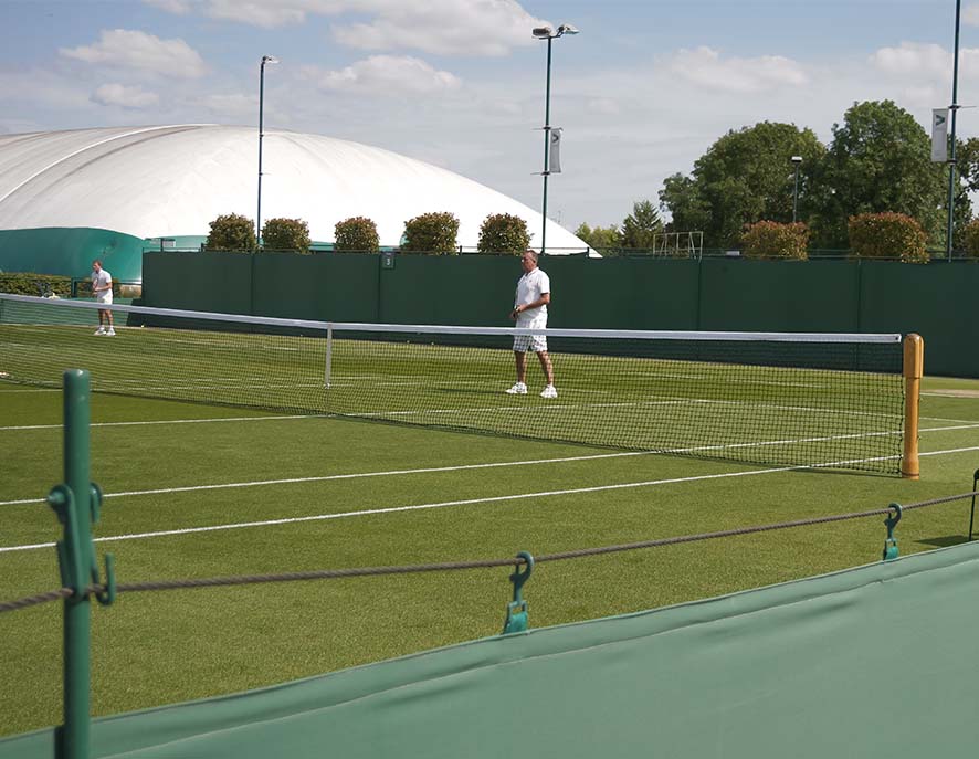 Two male tennis players in white playing on a green grass court with a domed sports complex in the background.