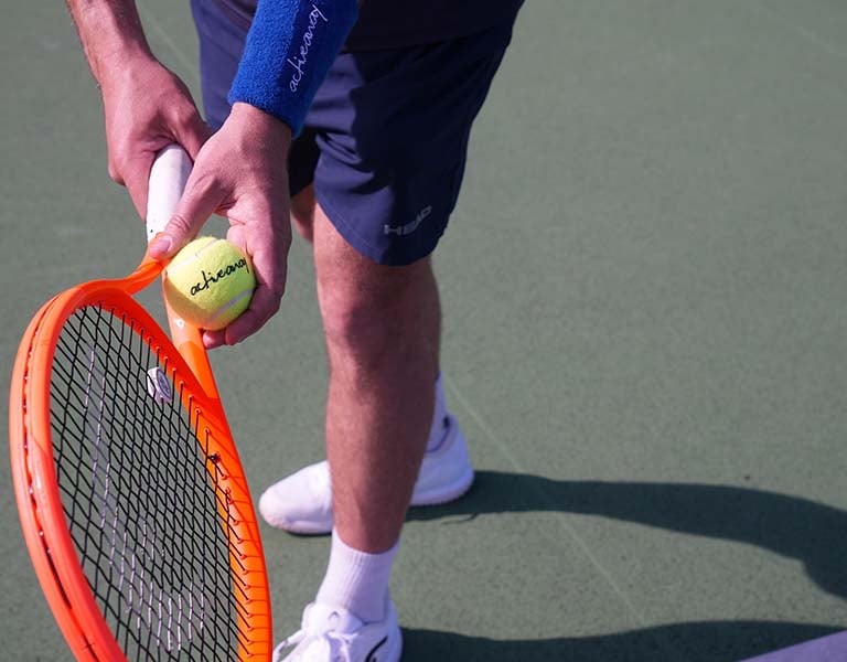 Tennis player holding a yellow tennis ball and an orange racket, ready to serve at the Active Away Junior Tennis Camps, Royal Russell School.