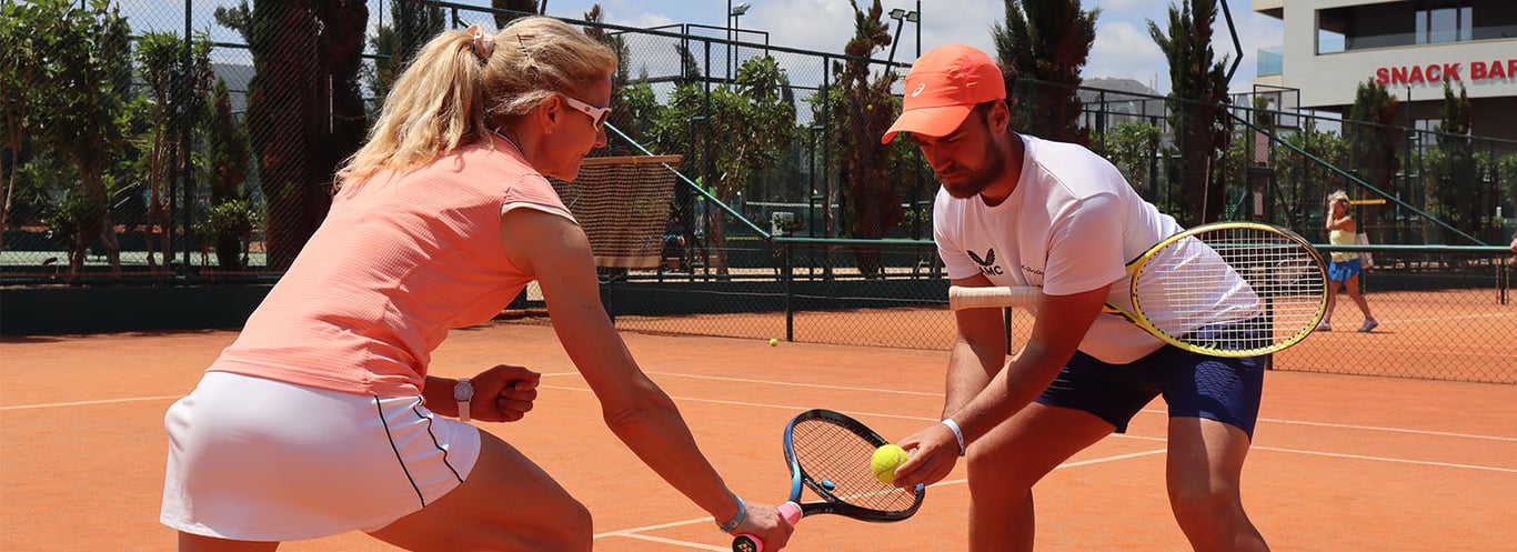 Tennis player receives coaching on a clay court from an instructor, both holding rackets, with tennis balls and 'Snack Bar' sign in the background.