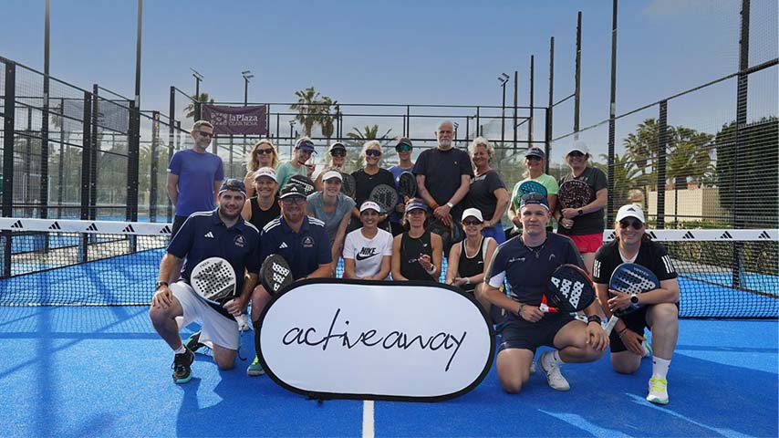 Group of people on a padel court under sunny skies holding rackets and an 'active away' banner, enjoying a padel holiday event.
