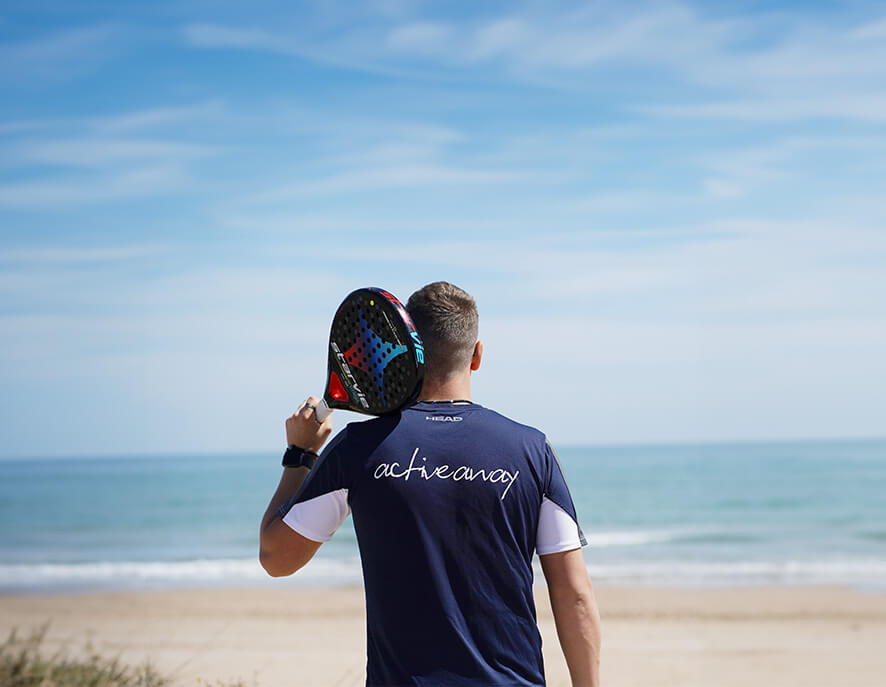 Man holding a paddle tennis racket on a beach, wearing an 'activeaway' shirt, looking towards the sea.