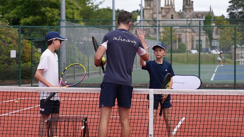 Tennis coach from Active Away instructing two young boys on a clay court with a historic building in the background.