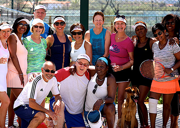 Group of adults in tennis attire posing on a court during a tennis holiday, with a dog sitting in front.