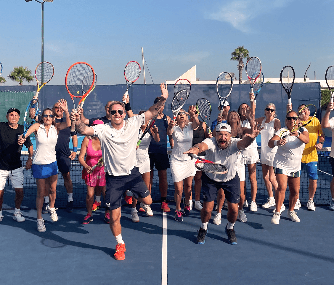 Group of adults in sporty tennis attire on a blue tennis court, smiling and holding rackets, with a sunny background and palm trees.