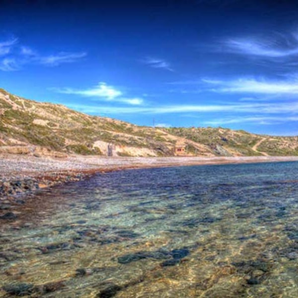 Aphrodite Hills Beach with clear waters and a rocky shoreline, set against a backdrop of gentle hills under a blue sky.
