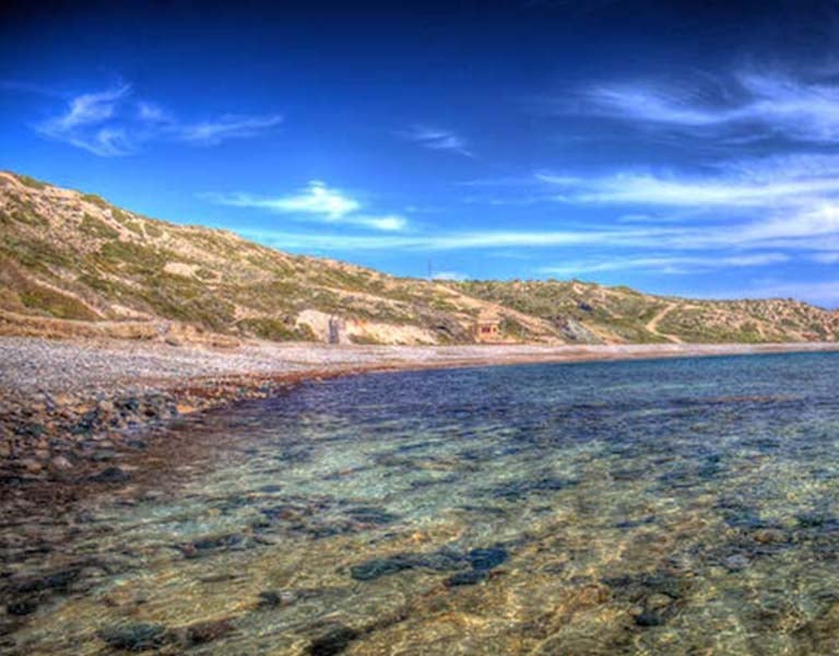 Aphrodite Hills Beach with clear waters and a rocky shoreline, set against a backdrop of gentle hills under a blue sky.