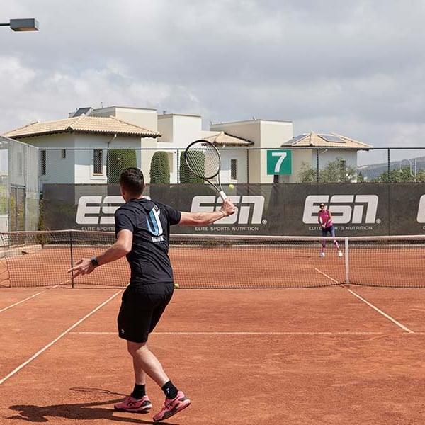 Tennis match on a clay court at Aphrodite Hills Resort with two players in action, surrounded by Mediterranean-style buildings and Elite Sports Nutrition branding.