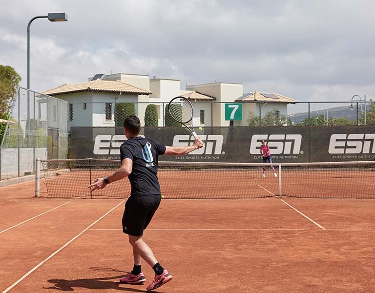 Tennis match on a clay court at Aphrodite Hills Resort with two players in action, surrounded by Mediterranean-style buildings and Elite Sports Nutrition branding.