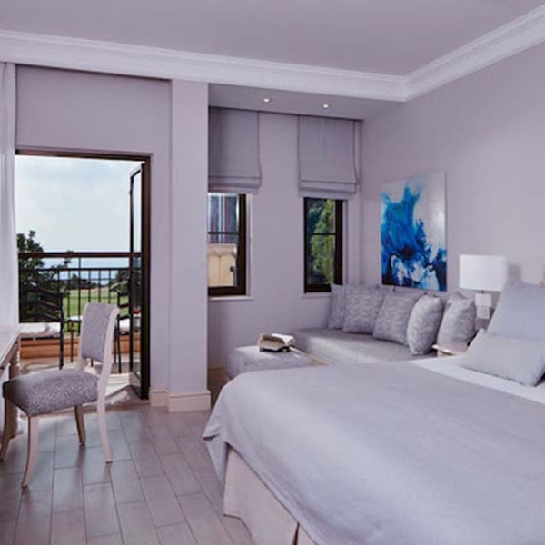 Deluxe Double Room at Aphrodite Hills with grey bedding, seating area, balcony view of greenery and sea.