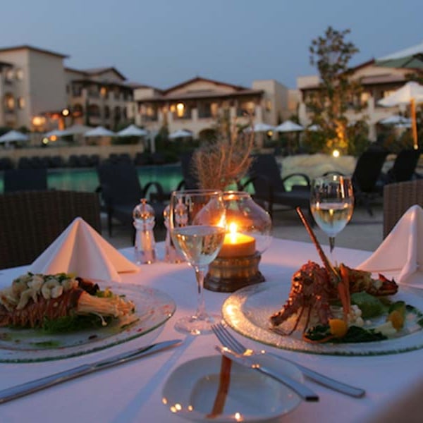 A beautifully set dining table for two with gourmet meals and a candle, overlooking a luxurious resort pool at Aphrodite Hills.