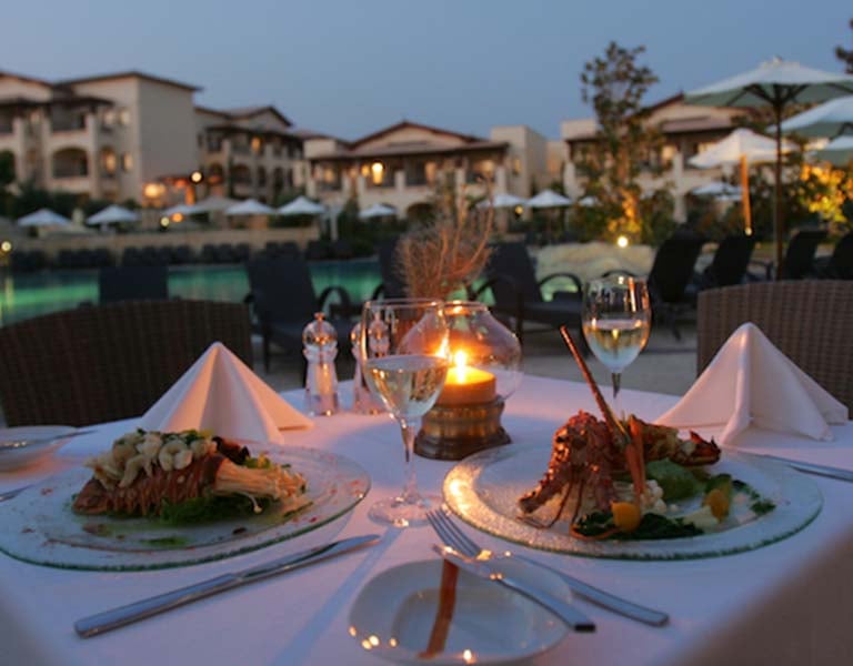 A beautifully set dining table for two with gourmet meals and a candle, overlooking a luxurious resort pool at Aphrodite Hills.