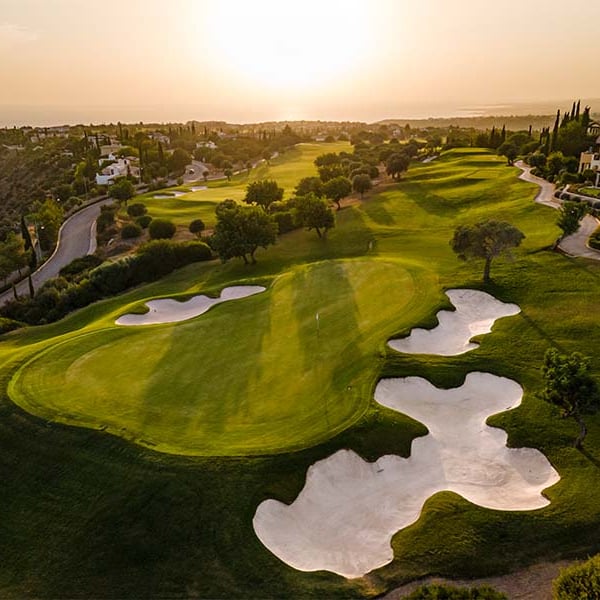 Aerial view of Aphrodite Hills Golf Course in Cyprus with green fairways, bunkers, sunset, and scenic surrounding landscapes.