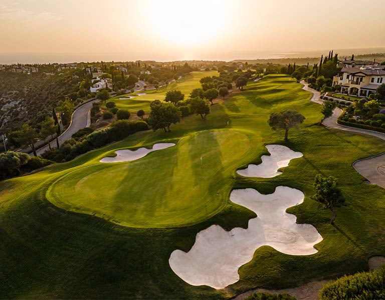 Aerial view of Aphrodite Hills Golf Course in Cyprus with green fairways, bunkers, sunset, and scenic surrounding landscapes.