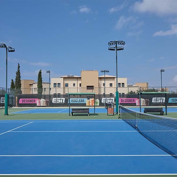 Hard tennis court at Aphrodite Hills with blue surface and advertisements, set against a modern building and partly cloudy sky.