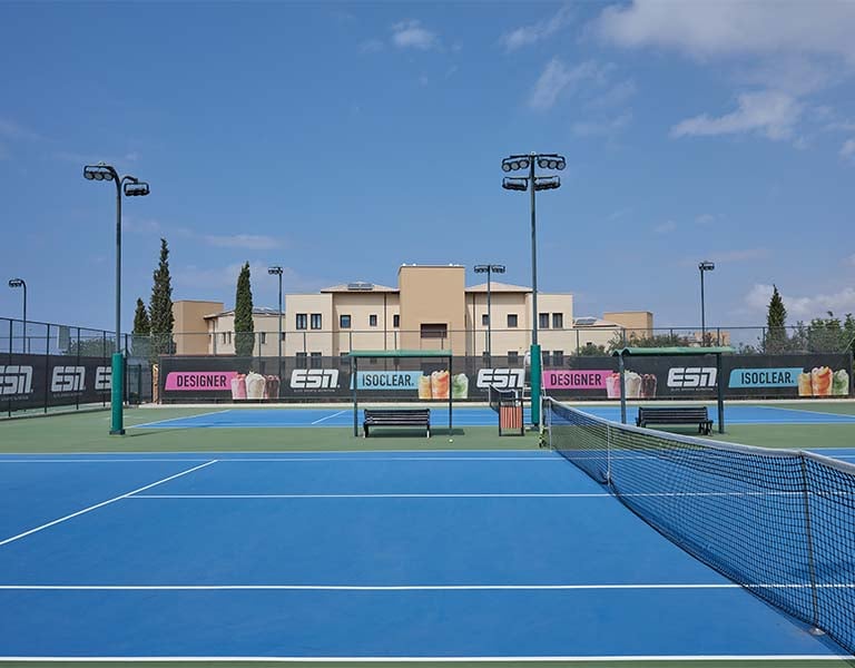 Hard tennis court at Aphrodite Hills with blue surface and advertisements, set against a modern building and partly cloudy sky.
