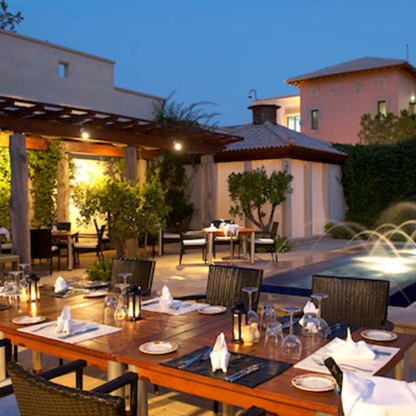 Outdoor dining area of Aphrodite Hills Resort's main restaurant in the evening, featuring elegantly set tables and a lit water feature.
