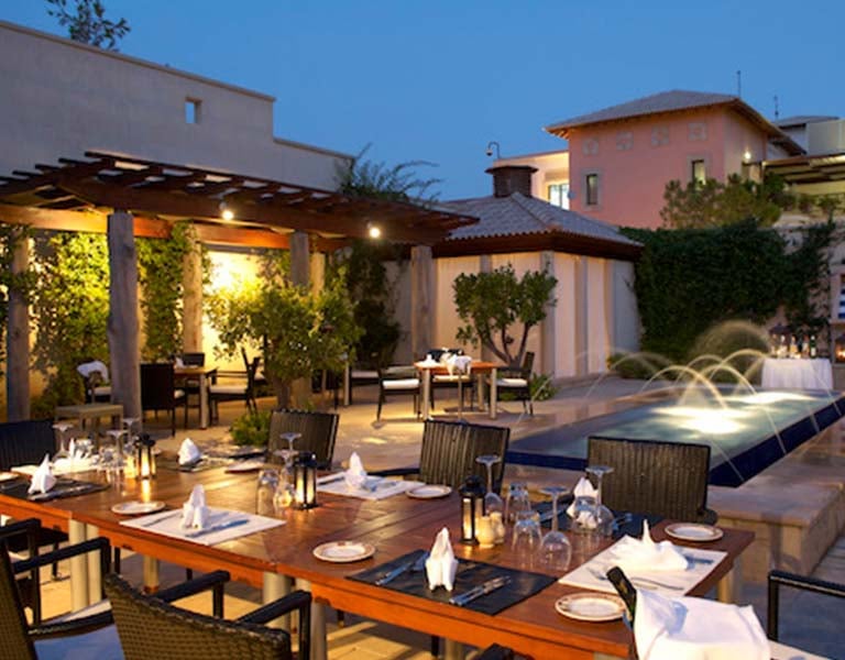 Outdoor dining area of Aphrodite Hills Resort's main restaurant in the evening, featuring elegantly set tables and a lit water feature.