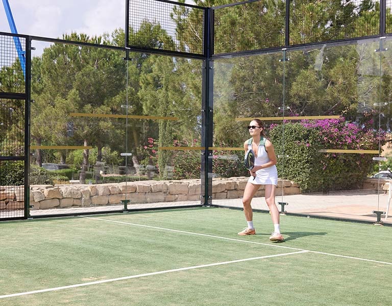 Woman playing padel tennis at Aphrodite Hills Resort, Cyprus, standing on the court with a racket and ready to receive a serve.
