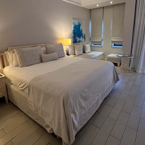 Hotel room at Aphrodite Hills with a double bed, white bedding, bedside lamps, decorative wall art, and large windows with grey curtains.