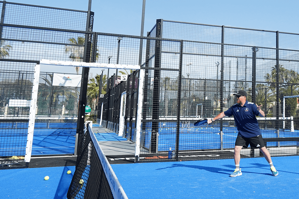 Man in navy sports shirt and cap ready to serve on a blue outdoor padel tennis court on a sunny day.