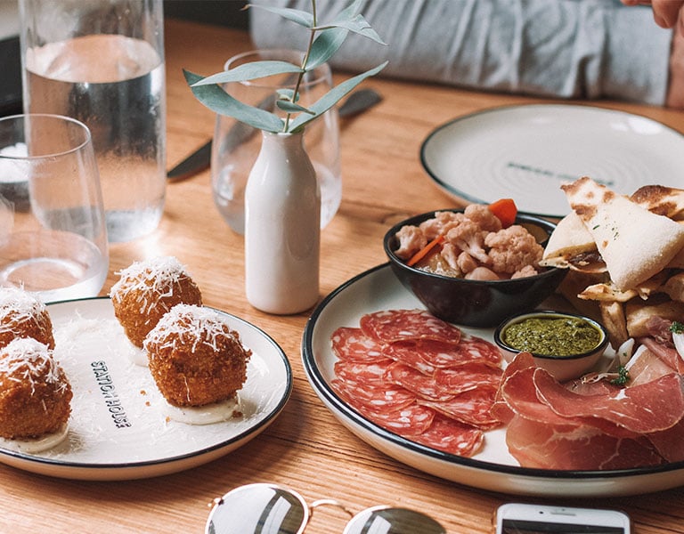 Assortment of Spanish tapas in Barcelona, featuring arancini balls, charcuterie platter with sliced meats, and pesto with pickled vegetables on a wooden table.