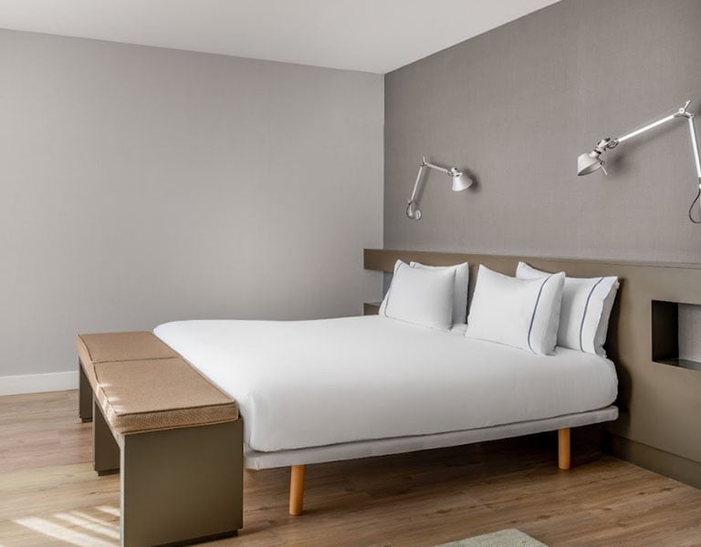 Minimalist hotel room in Barcelona with a double bed, white linens, wall-mounted lamps, and a sleek bench at the bed's foot.