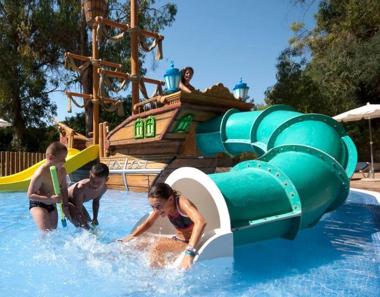 Children playing on a water slide at a beach club, with a pirate ship backdrop and a girl sliding into a shallow pool.