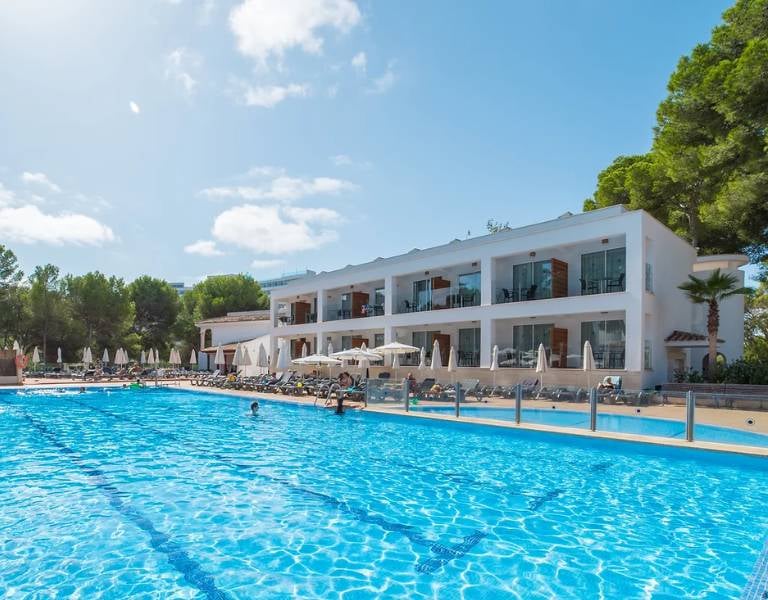 Outdoor pool area at a beach club with a clear blue pool, sun loungers, parasols, and a two-storey building surrounded by trees.