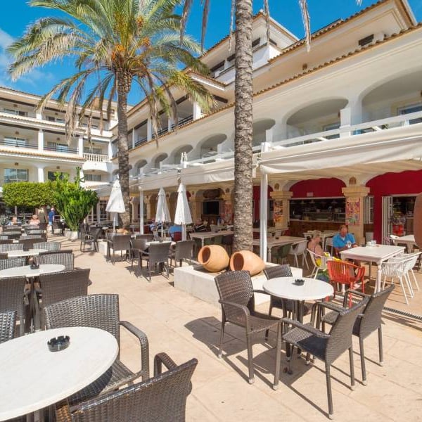 Outdoor dining area at a beach club with tables, chairs, white parasols, and a Mediterranean-style building in the background.