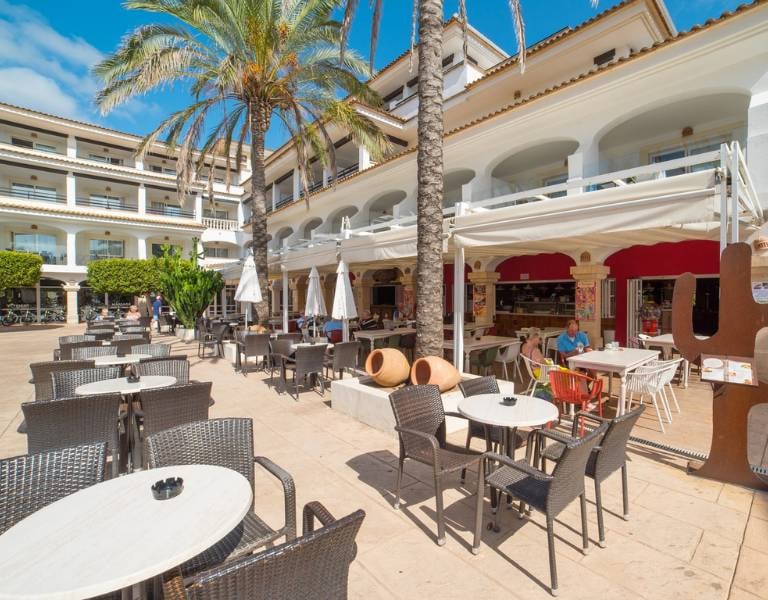 Outdoor dining area at a beach club with tables, chairs, white parasols, and a Mediterranean-style building in the background.