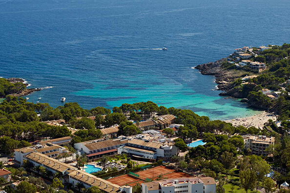 Aerial view of Beach Club Font de Sa Cala, Majorca, featuring resort buildings, tennis courts, and a blue sea surrounded by greenery.