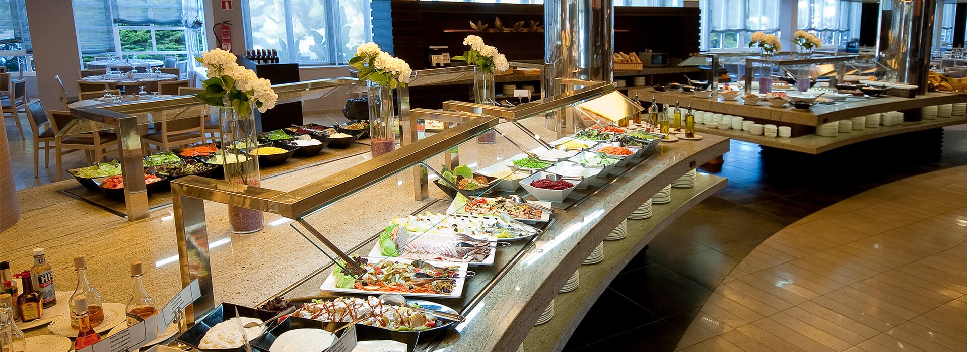 Buffet at a beach club displaying salads, seafood, and gourmet platters with flowers and natural light in a spacious dining area.