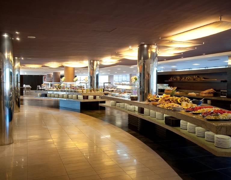 Luxurious beach club buffet with curved counters and an assortment of fruits, pastries, and gourmet dishes under ambient lighting.