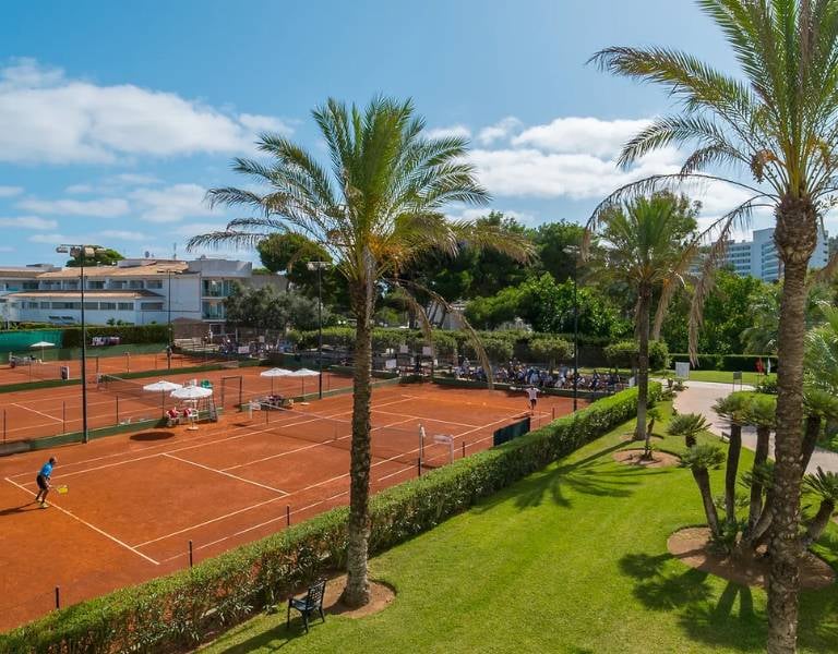Tennis courts at a beach club surrounded by greenery and palm trees, with players in action against a backdrop of club facilities and clear skies.