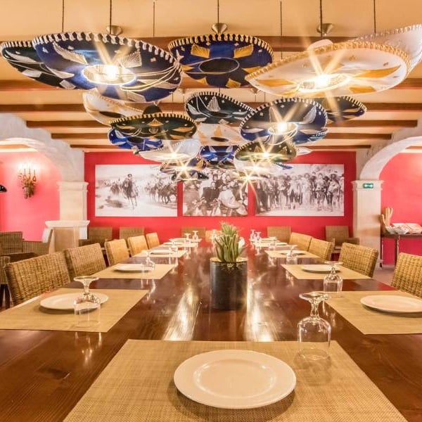 Elegant dining room at a beach club with a wooden table, woven placemats, white plates, and decorative umbrella lights, set against coral walls with a beach scene mural.