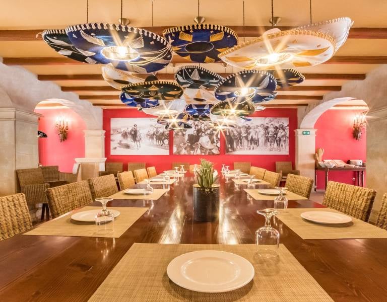 Elegant dining room at a beach club with a wooden table, woven placemats, white plates, and decorative umbrella lights, set against coral walls with a beach scene mural.