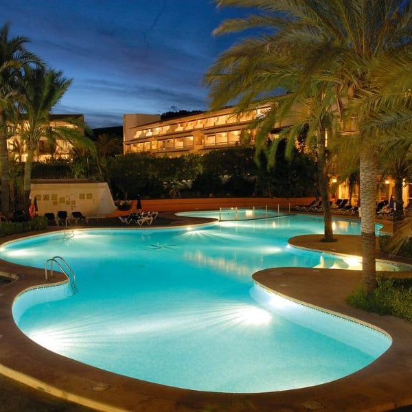 Night view of a beach club swimming pool surrounded by palm trees, with a hotel building illuminated in the background.