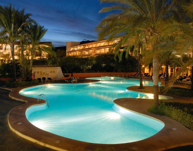 Night view of a beach club swimming pool surrounded by palm trees, with a hotel building illuminated in the background.