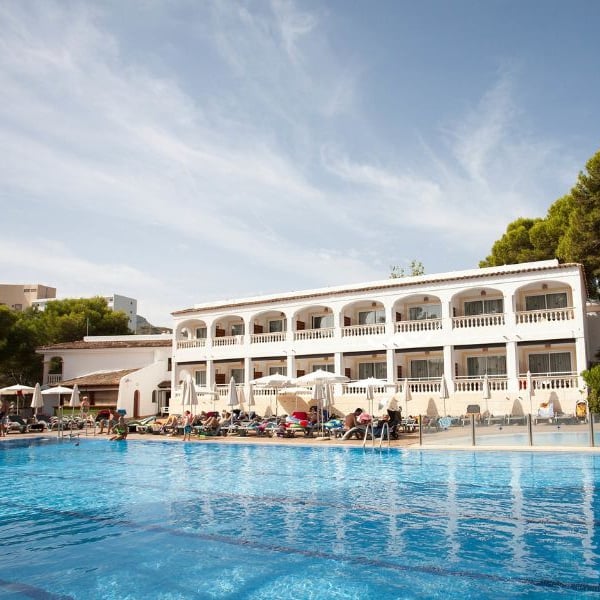 Outdoor pool at a beach club, with a Mediterranean-style building, surrounded by sunbathers and pine trees.